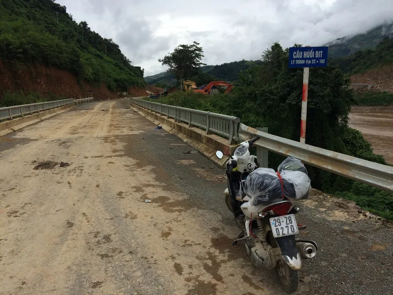 A muddy motorbike parked on the Cầu Huổi Dịt bridge amid misty mountains in Lai Châu province, Vietnam.