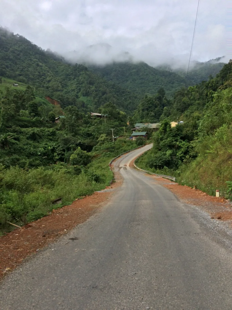 A winding mountain road passes small houses beneath mist-covered peaks in Lai Chau province, Vietnam.
