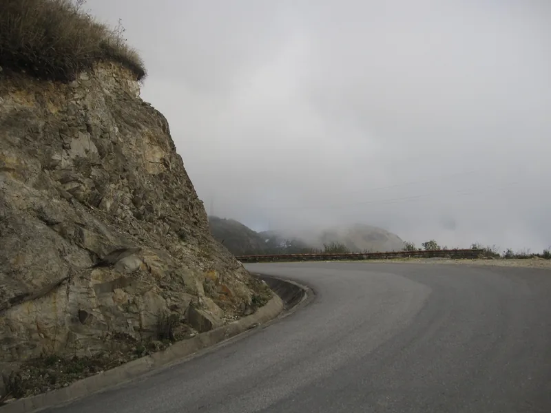 A curving mountain road with a rocky cliff wraps through thick fog on the O Quy Ho Pass in Lai Chau.