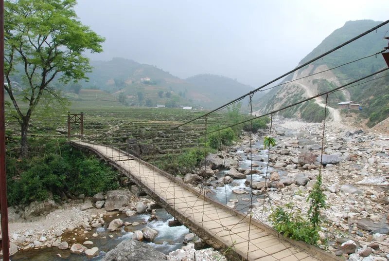 A cable suspension footbridge crosses a rocky river valley surrounded by terraced hills in Lai Chau province.