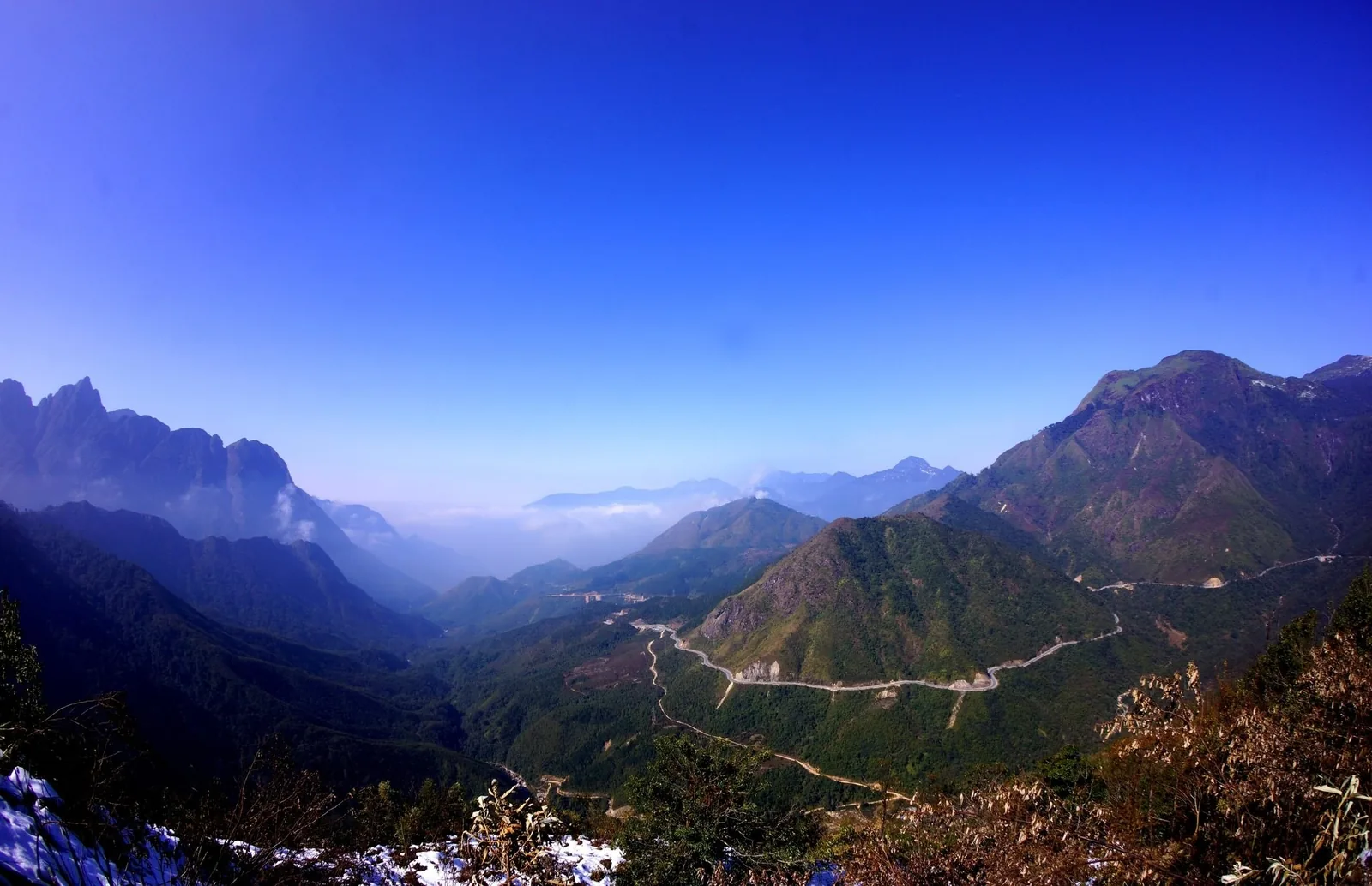 A winding mountain road snakes through green peaks under a clear blue sky at O Quy Ho Pass in Lai Chau.