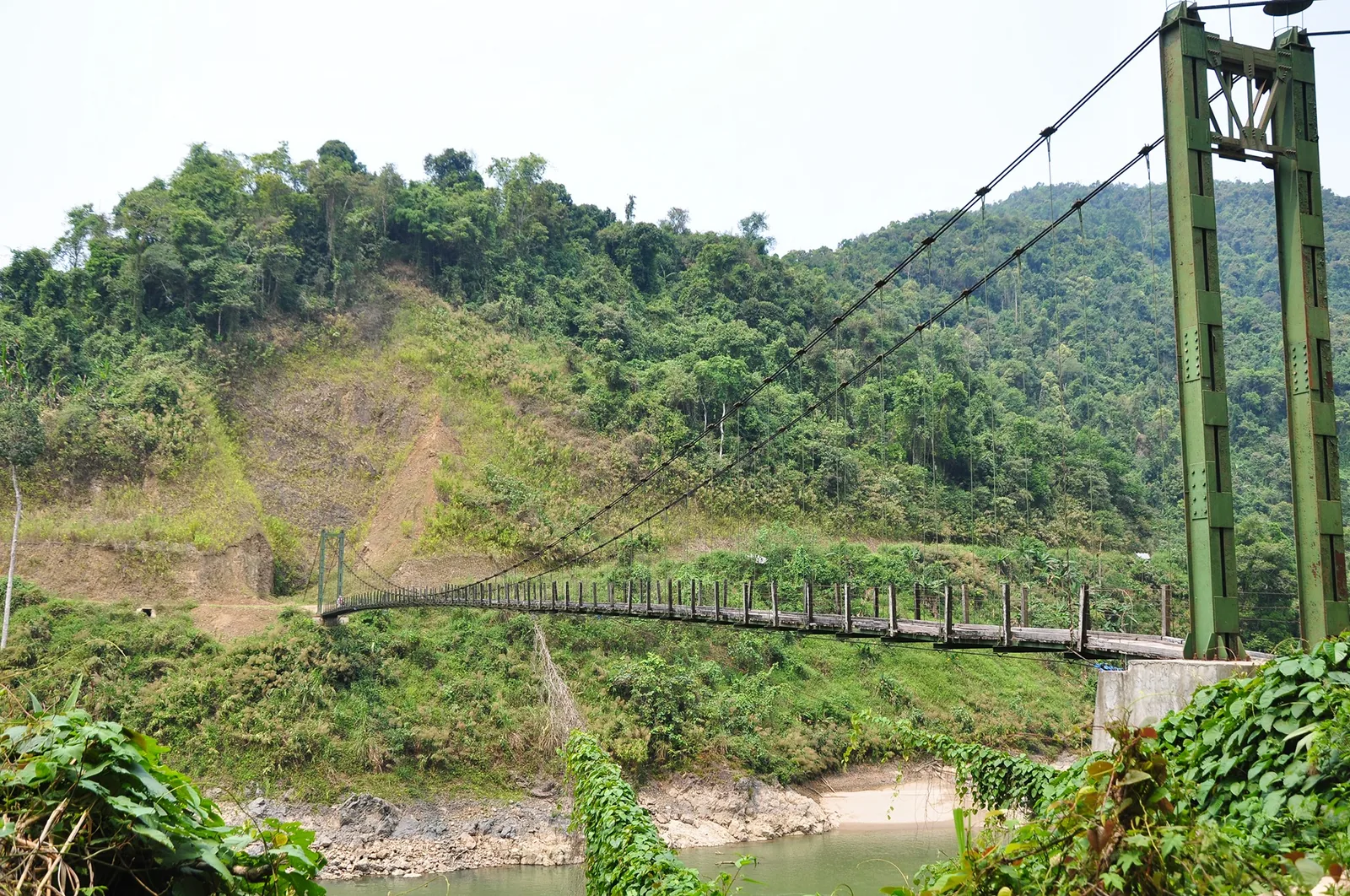 A green suspension bridge crosses a river surrounded by forested hills near the upper Da River in Lai Chau.