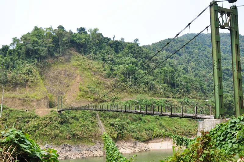 A green suspension bridge crosses a river surrounded by forested hills near the upper Da River in Lai Chau.